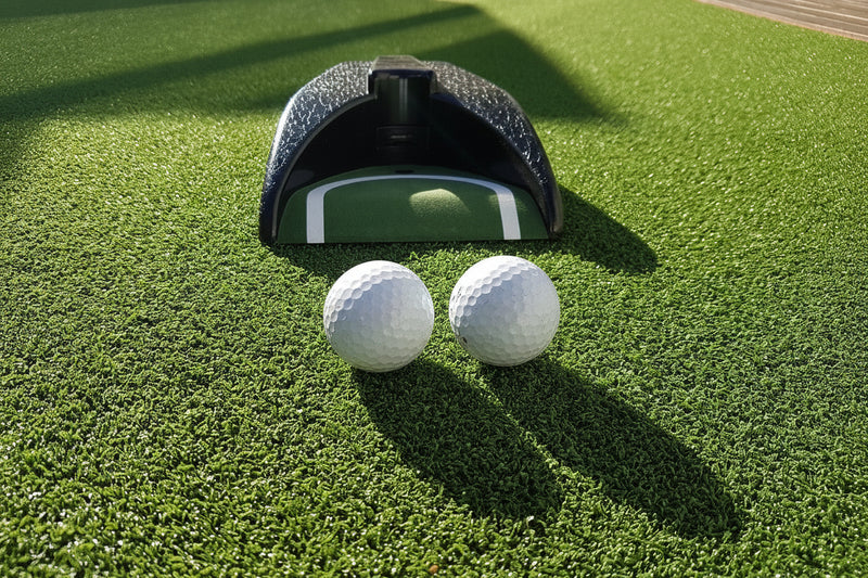 Two golf balls on a green surface with a golf ball return device in the background.
